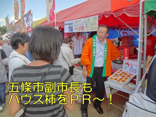 五條市副市長もハウス柿をPR。屋台の前で、オレンジのはっぴを着た五條市副市長が柿をお客さんへPRしている写真