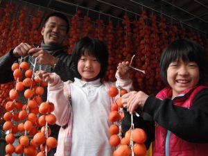 紐で繋いだ柿を吊るすお手伝いをしている笑顔の子供たちの写真