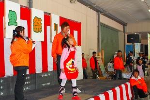 舞台の上オレンジのジャンバーを着たスタッフ2人と女の子1人の写真