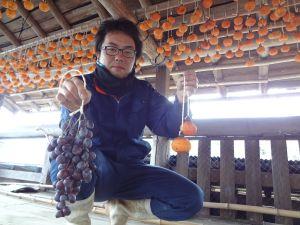 屋根いっぱいに柿を吊るした倉庫の下で糸にくくったぶどうを片手にもつ生産者の写真
