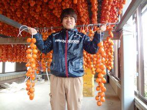 20個ほどの皮をむいた柿を左右交互に紐で吊るしたものを、両手に持っている生産者の写真