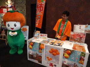 奈良まほろば館にてマスコットキャラクターのカッキーと一緒にハウス柿のPRを行っている様子の写真