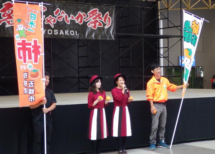 ステージ前で、柿を持ちながらマイクで話している女性二人と、その両脇でのぼりを持つ男性スタッフの写真
