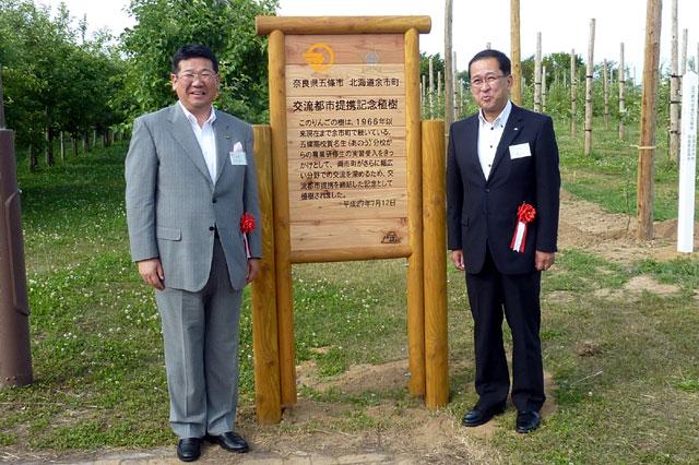 記念植樹を行いました 緑の中の看板の前で、嶋余市町長と太田五條市長が並んでいる写真