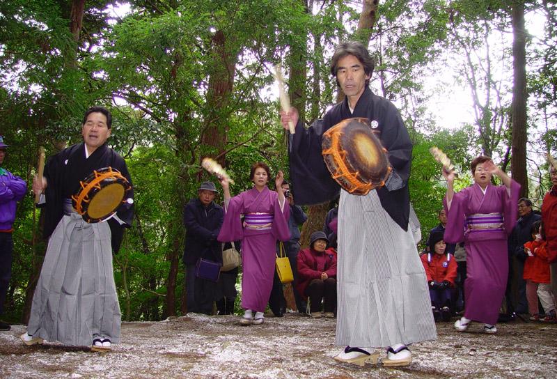 袴を着た男性が太鼓を打ちその後方で紫色の着物を着た女性が踊る写真