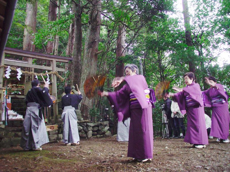 鳥居の前で袴を着た男性が太鼓を打ちその後方で紫色の着物を着た女性が踊る写真